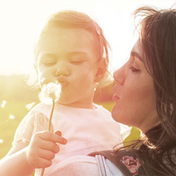 mother holding child while child blows on dandilion